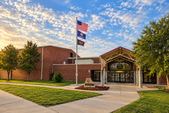 Exterior view of a brick school building with a flagpole displaying the American flag, Kansas flag, and a black-and-red flag in front of the glass entrance.